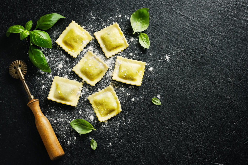 Italienische Ravioli mit Mehl bestäubt liegen neben Basilikumblättern und Ravioli-Küchenwerkzeug