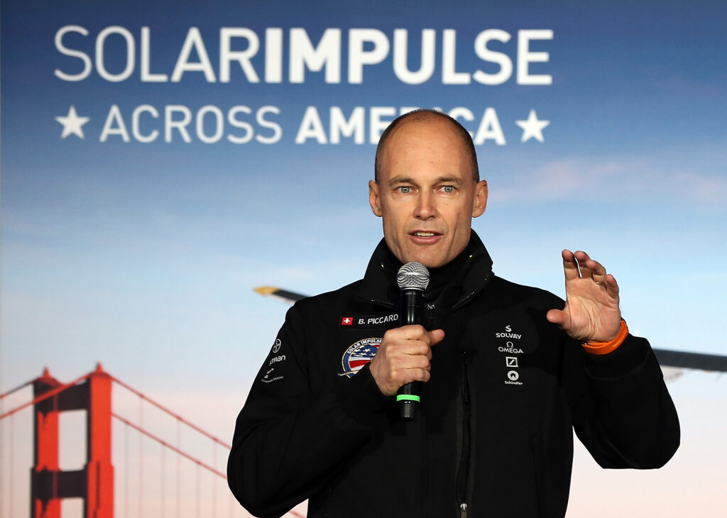 Bertrand Piccard hält ein Mikrofon in der Hand und hebt eine Hand