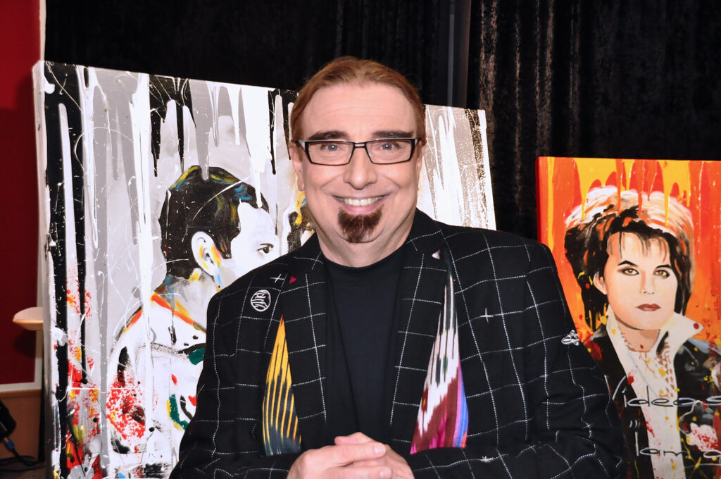 Rudi Dolezahl steht mit schwarz-weiß kariertem Sakko und buntem Schal direkt vor der Kamera und lächelt. Er trägt eine Brille. Hinter ihm stehen zwei sehr bunte Bilder von zwei Menschen.