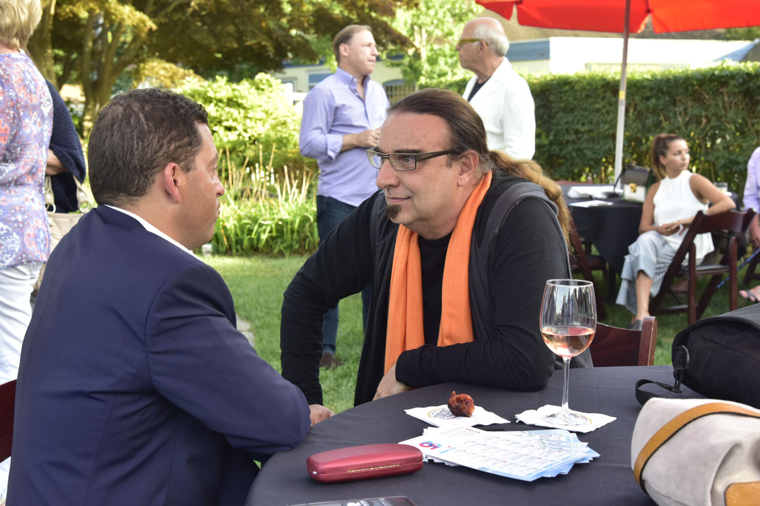 Rudi Dolezal mit schwarzem Hemd und organgem Schal sitzt an einem Tisch auf dem Unterlagen liegen und ein Glas Wein steht. Er gibt einem Mann in blauem Anzug der ihm gegenübersitzt ein Interview