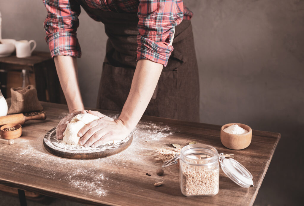 Bäcker formt Brot Bäcker mit kariertem Hemd hat beide Hände auf einem Brot-Teigling. Daneben steht ein Behälter mit Salz und Getreide
