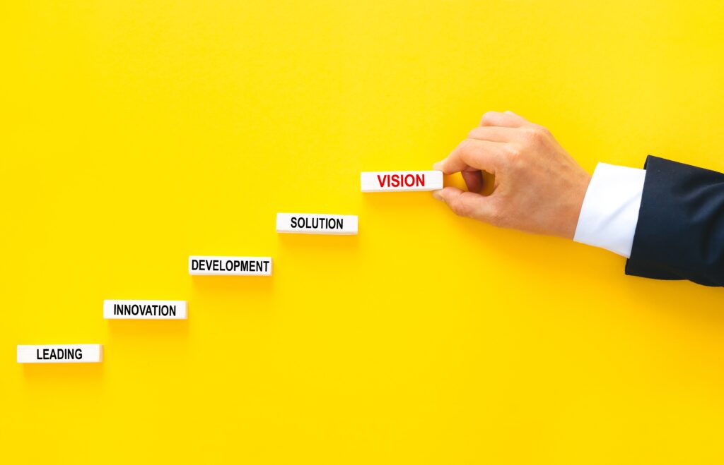 Gelber Hintergrund auf dem Leading, Innovation, Development, Solution und Vision in Bausteinen wie eine Stiege aufgebaut werden. Das letzte Wort Vision hält eine Hand mit weißer Bluse und dunklem Anzug