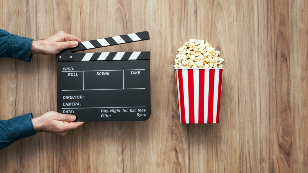 Brauner Holzhintergrund. Davor zwei Hände die eine Filmklappe halten. Daneben eine rot-weiß gestreifte Tüte mit Popcorn