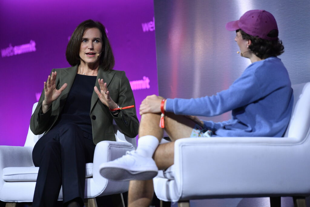 Judith Wiese Judith Wiese sitzt auf einer Bühne und gestikuliert mit ihren Händen. Neben ihr ein Mann in blauem Sweatshirt und violetter Kappe mit kurzer Hose, weißen Tennissocken und weißen Turnschuhen, der in einem weißen Sessel sitzt.