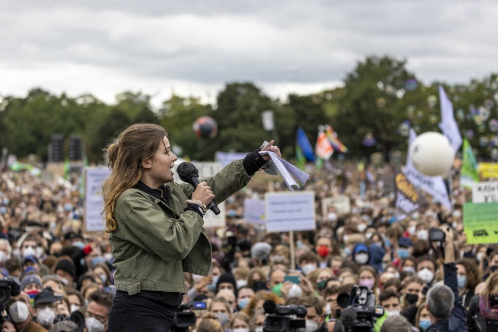 Luisa Neubauer Luisa Neubauer ist von der Seite abgeildet. Sie hält ein Mikrofon und ein paar Zettel in der Hand und spricht zu unzähligen Menschen von einer Bühne