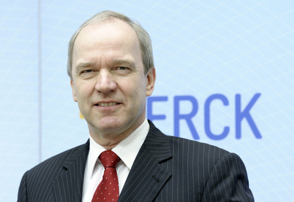 Karl-Ludwig Kley trägt als Direktor der MERCK Gruppe einen gestreiften Anzug und eine rote Krawatte mit weißen Punkten. Er blickt in die Kamera. Hinter ihm das Logo von MERCK