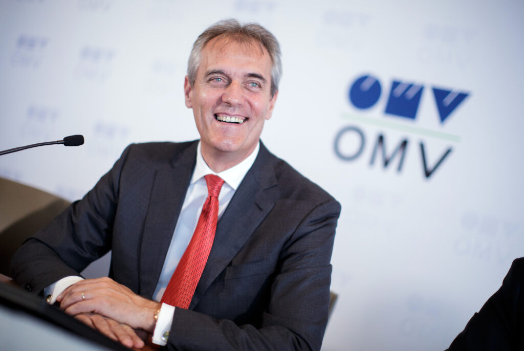 Rainer Seele Rainer Seele mit dunklem Anzug und roter Krawatte lacht in die Kamera. Hinter ihm das Logo der OMV
