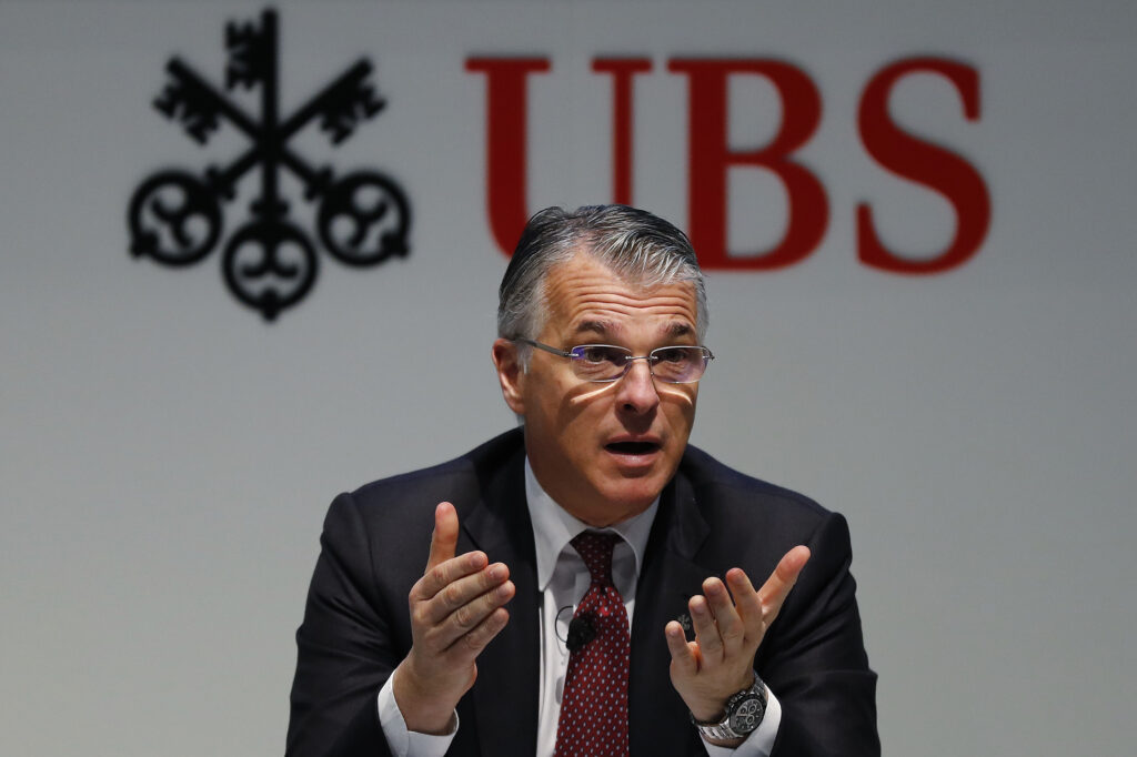 Sergio Ermotti Sergio Ermotti trägt eine Brille und hält die Hände offen in die Kamera. Hinter ihm das Logo von UBS