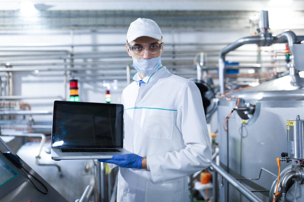 Mann mit Mundschutz und weißer Hygienekleidung sowie blauen Hygienehandschuhen trägt eine Brille und hält in einen Computer in die Kamera