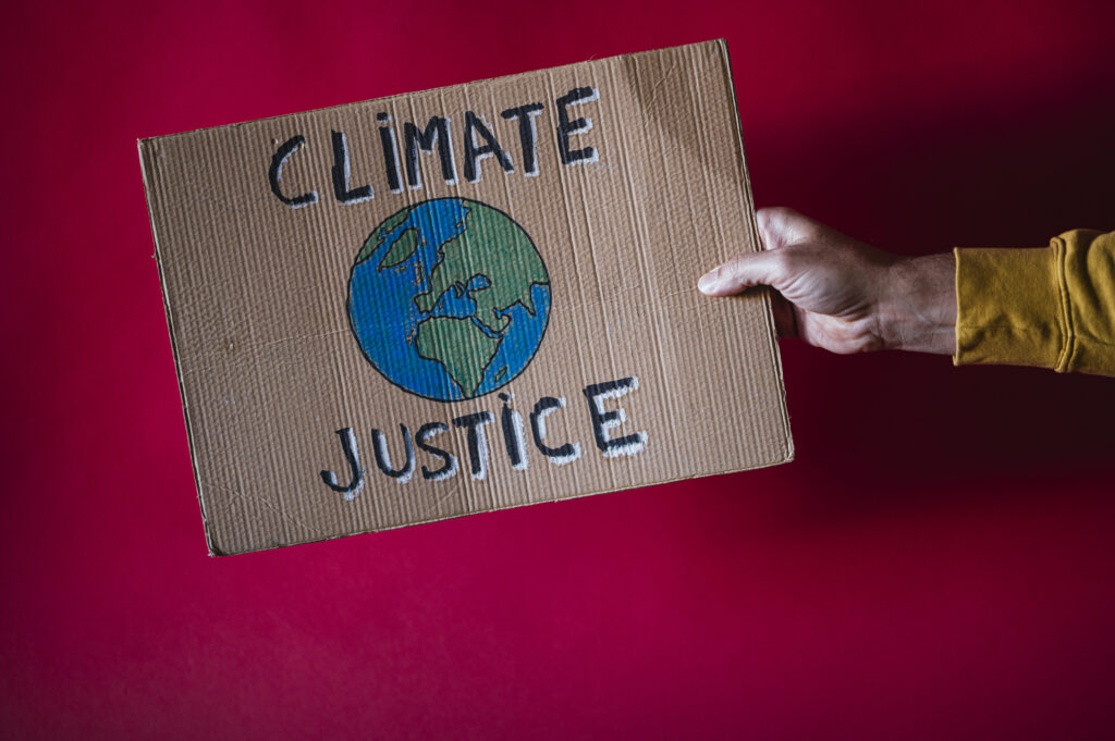 Luisa Neubauer Roter Hintergrund vor dem eine Hand ein Plakat aus Karton mit der Aufschrift CLIMATE JUSTICE hält. In der Mitte ist die Erdkugel abgebildet