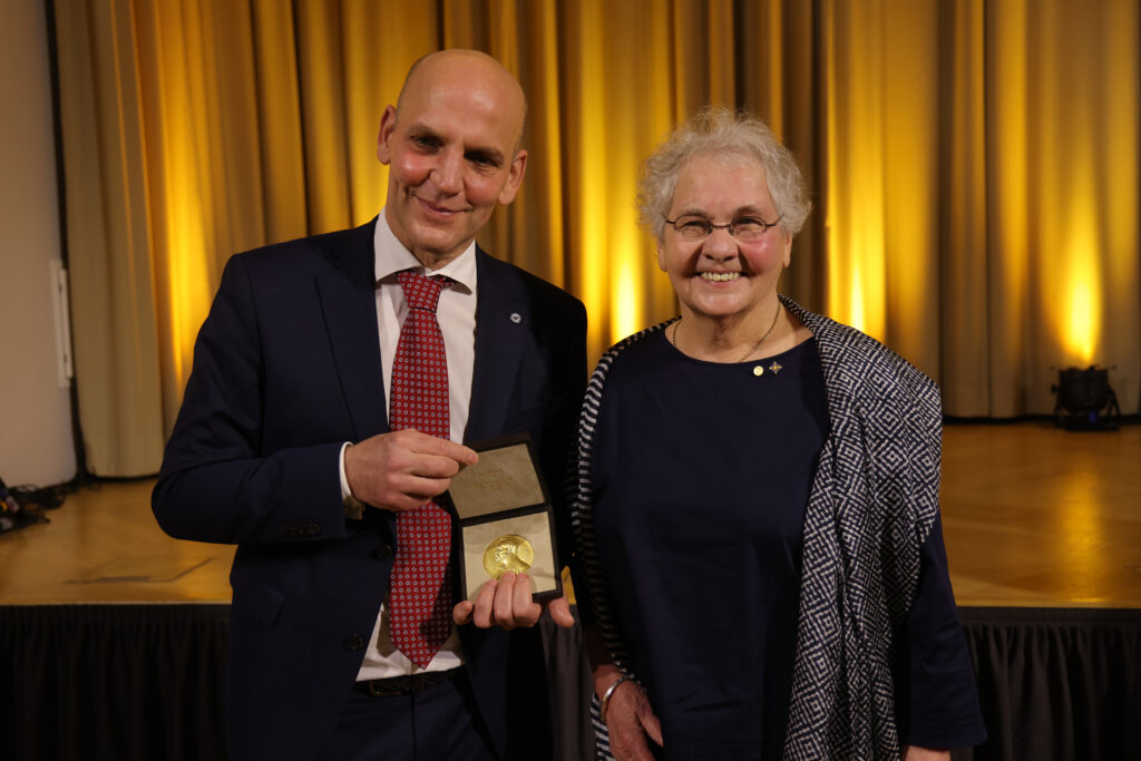 Nüsslein-Volhard Christiane Nüsslein-Volhard steht mit einem Mann, der den Nobelpreis hält vor einer Bühne und lächelt in die Kamera