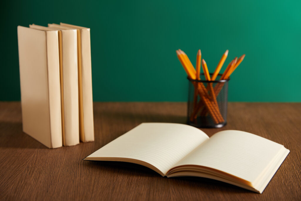 Marcel Eris Ein aufgeschlagenes Buch mit leeren Seiten. Daneben links weitere drei Bücher und ein Behälter der Bleistifte beinhaltet. Das Buch liegt auf einem braunen Tisch