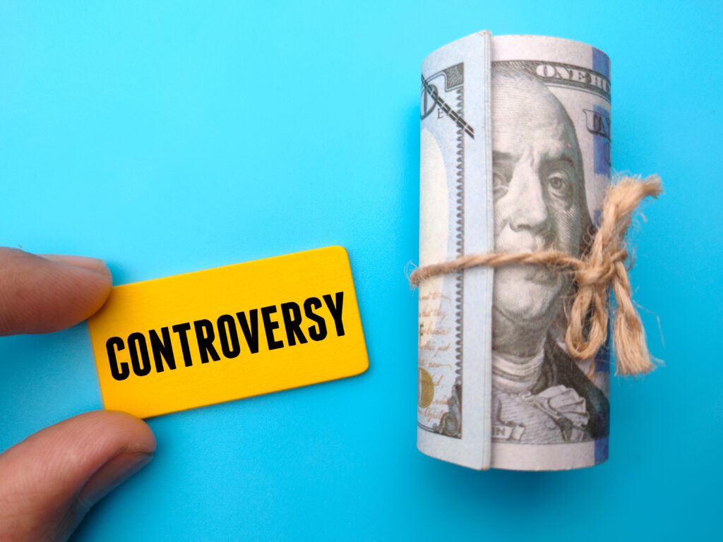 Marcel Eris Blauer Hintergrund auf dem eine Hand ein gelbes Schild hält, auf dem Controversy steht. daneben ein Bündel mit Banknoten die mit einer Schnur umgarnt sind
