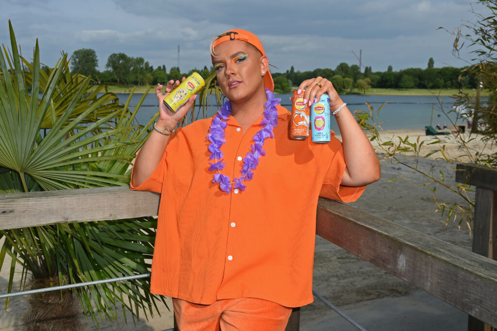 twenty4tim twenty4tim stark geschminkt mit oranger Bluse und oranger Kappe sowie einer lila Blumenkette um den Hals hältdrei Lipton-Eistee Dosen in seinen Händen.
