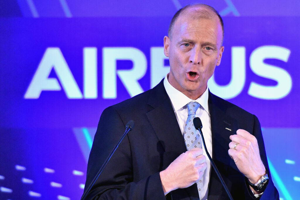 Tom Enders trägt einen dunklen Anzug und eine blaue Krawatte mit Punkten. Er steht auf einer Bühne mit Mikrofon. Dahinter das Logo von Airbus