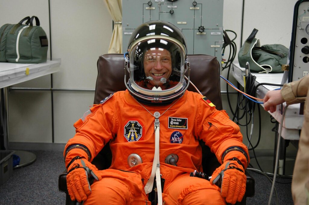 Thomas Reiter Thomas Reiter sitzt in einem orangen Raumanzug und schwarzem Helm in einem Sessel und lächelt in die Kamera
