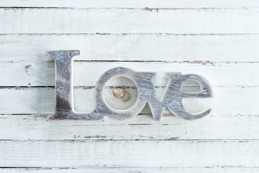 twenty4tim weißer Hintergrund aus Holz. Darauf die Lettern Love geschrieben. Im O liegen zwei Eheringe
