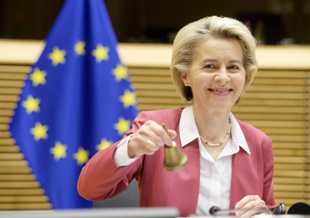Ursula von der Leyen Ursula von der Leyen mit rosa Sakko und weißer Bluse hält eine goldene Glocke in der Hand. Hinter ihr die EU-Flagge. Sie sitzt an einem Tisch