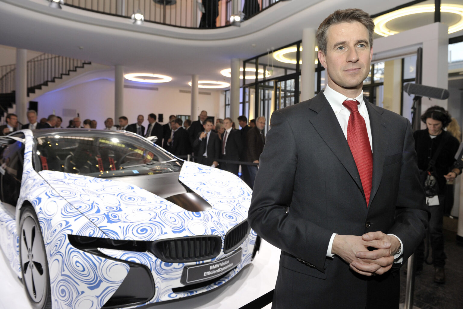 Stefan Quandt steht mit Anzug und roter Krawatte vor einem modernen neune BWM im futuristischem Design