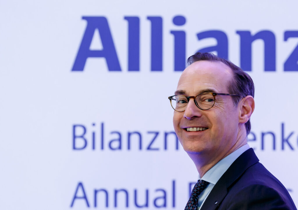 Oliver Bäte lächelt seitlich in die Kamera. Hinter ihm das Logo der Allianz