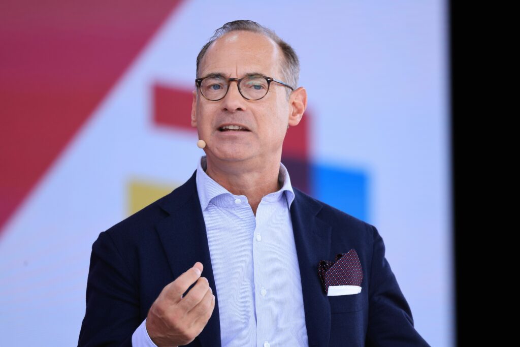 Oliver Bäte Oliver Bäte trägt einen Anzug, eine Brille und ein blaues Hemd. Um den Hals hat er ein kleines Mikrofon umgehängt. Er blickt seitlich in die Kamera