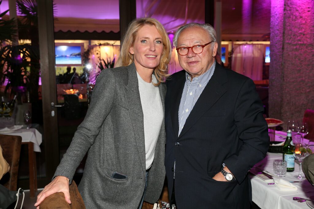 Hubert Burda mit Anzug steht neben seiner Ehefrau und lacht in die Kamera. Er trägt eine rote Brille