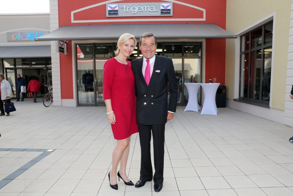 Wolfgang Grupp in dunklem Anzug steht neben seiner Gattin, die ein elegantes kurzes rotes Kleid trägt vor dem Eingang zu einem Geschäft von Trigema