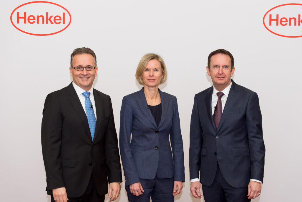 Casrten Knobel Carsten Knobel mit zwei weiteren Personen in Anzügen vor dem Logo von Henkel