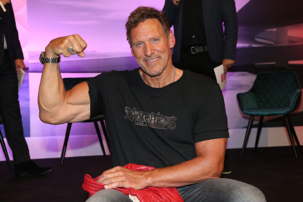 Ralf Möller sitzt in einem schwarzen T-Shirt und hält seinen rechten Arm hoch wo der seine Muskeln zeigt