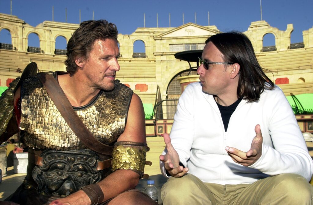 Ralf Möller Ralf Möller sitzt in der Rolle als Gladiator neben seinem Regisseur in deiner Kulisse, die eine Arena zeigt im Hintergrund