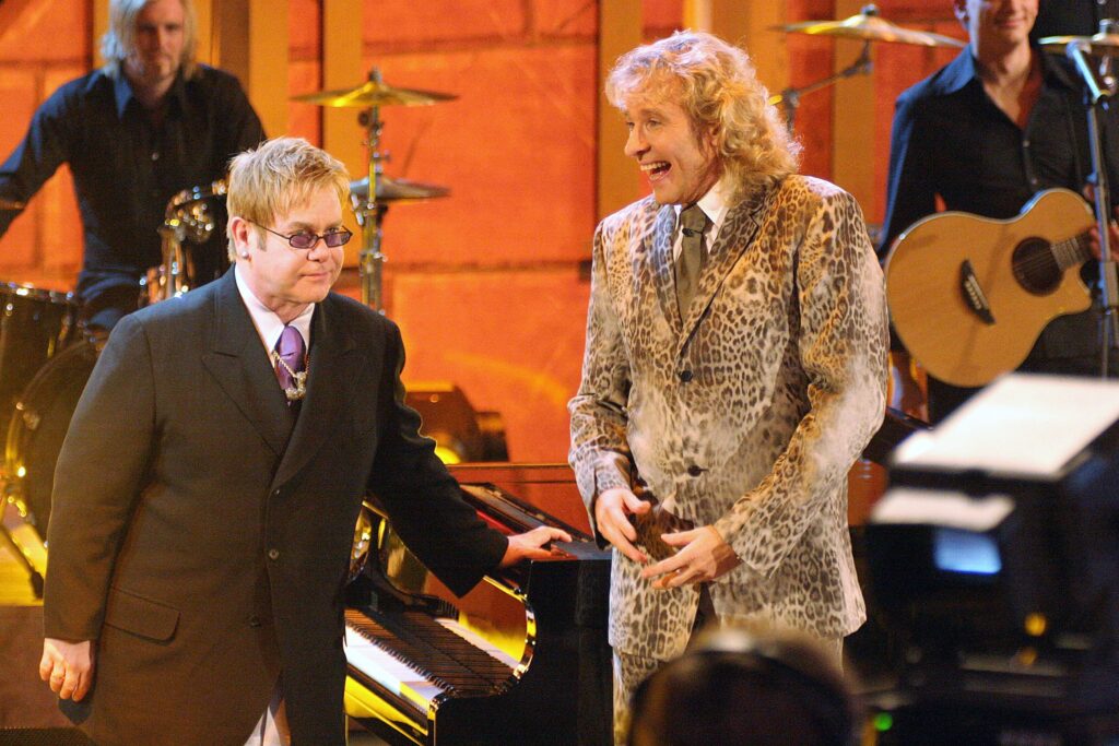 Thomas Gottschalk Thomas Gottschalk trägt einen Anzug in Leopardenoptik und steht neben Elton John, der einen schwarzen Anzug und eine dunkle Brille trägt. Dahinter Musiker
