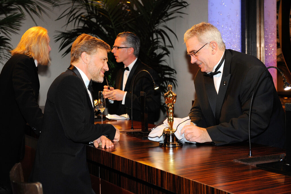Christoph Waltz Christoph Waltz steht an einer Bar einem anderen Mann mit schwarzem Anzug gegenüber. Vor den beiden eine Oscar-Trophäe am Tisch