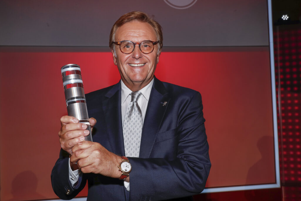 Roland Mack steht vor einer roten Wand und hält einen Preis in seinen Händen