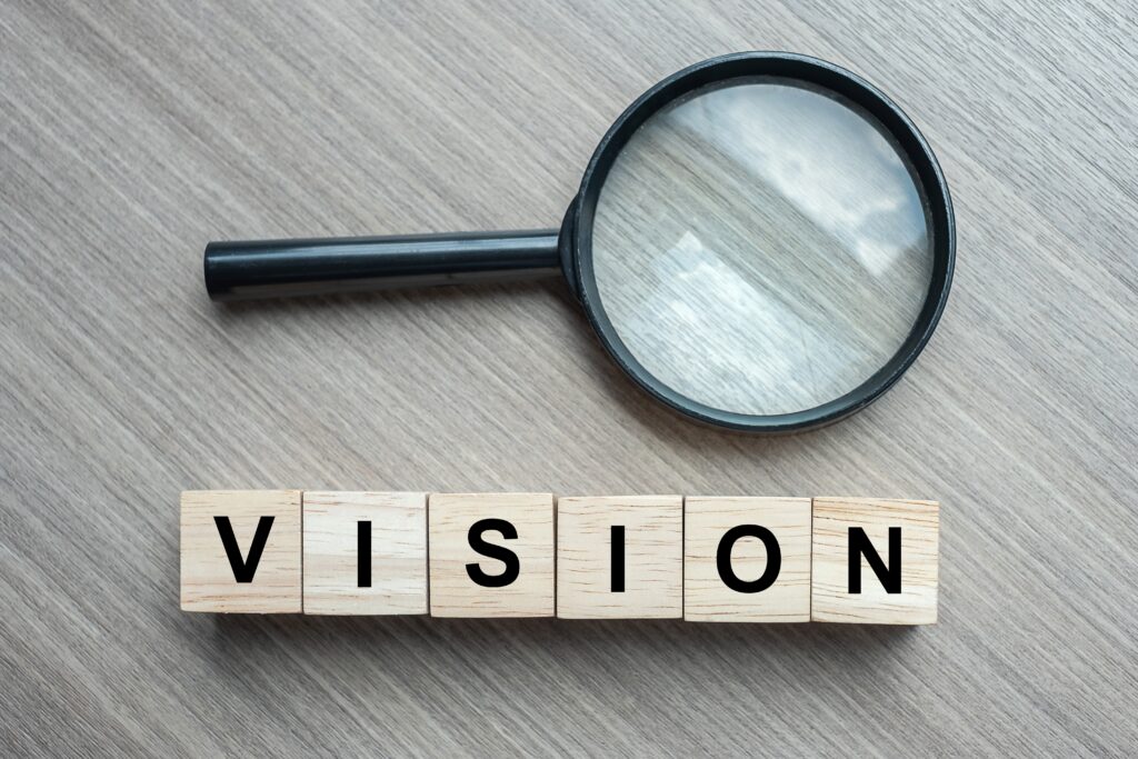 Helle Holzplatte auf der das Wort Vision in Großbuchstaben auf Würfeln zu lesen ist. Dahinter eine schwarze Lupe