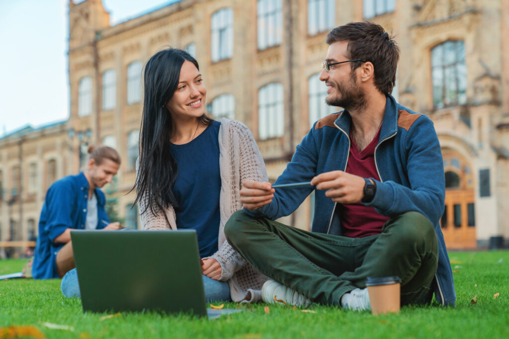 Hasso Plattner Eine Mann und eine Frau sitzen vor einer Universität im Gras. Die Frau hat einen Computer vor sich aufgestellt und lächelt den Mann an. Er sitzt im Schneidersitz