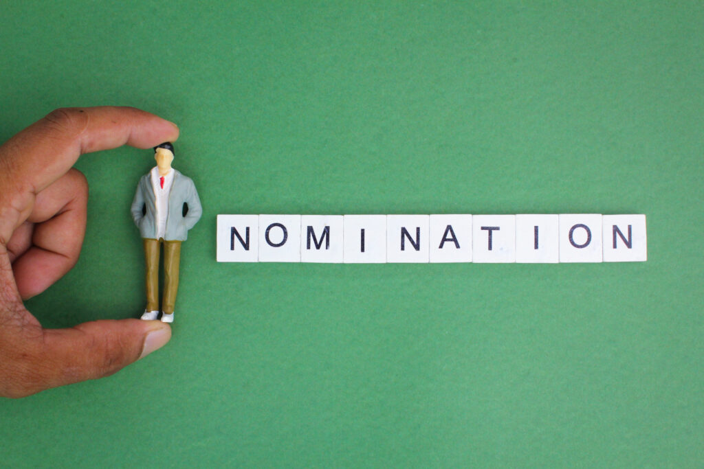 grüner Hintergrund mit einer Hand die eine Plastikfigur hält. Daneben die Buchstaben: Nomination
