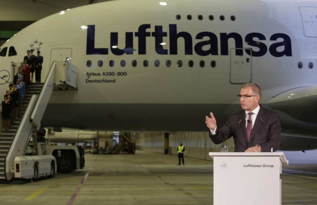 Carsten Spohr Carsten Spohr an einem Rednerpult hebt eine Hand in die Höhe. Dahinter eine Maschine der Lufthansa