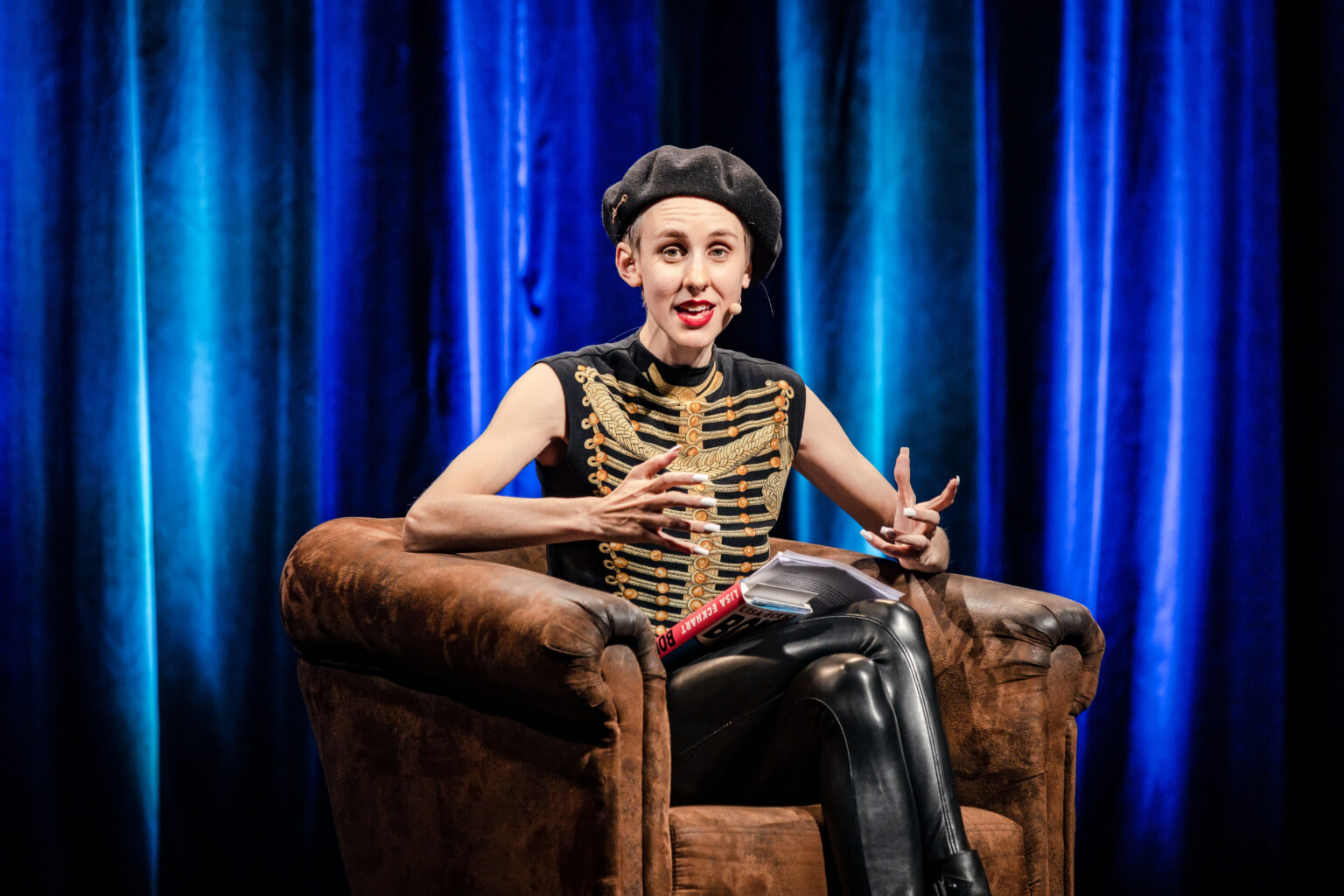 Lisa Eckhart sitzt mit einer schwarzen Kappe und einer Lederhose in einem braunen Sofa auf einer Bühne