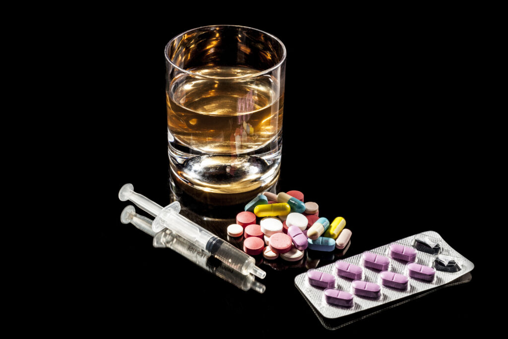 Alkohol im Glas, eine Spritze und viele Tabletten liegen auf einem schwarzen Hintergrund