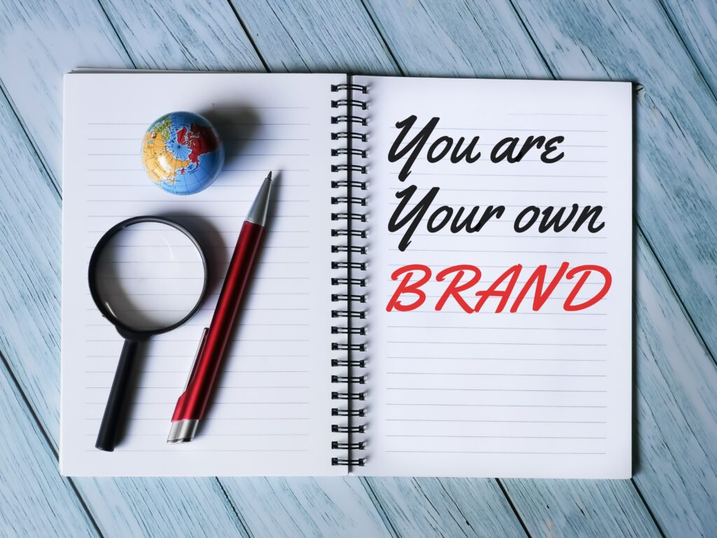 Buch mit einer Lupe, einer Weltkugel und einem Kugelschreiber und auf der anderen Seite steht: you are your own Brand