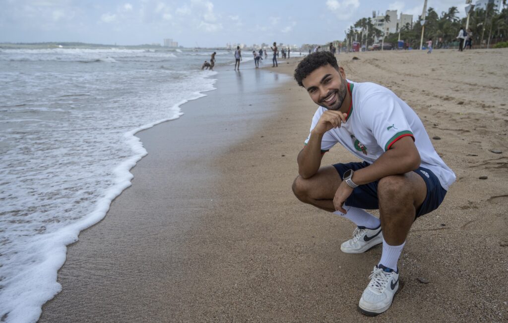 Younes Zarou mit T-Shirt und kurzer Hose hockt an einem Strand und blickt in die Kamera