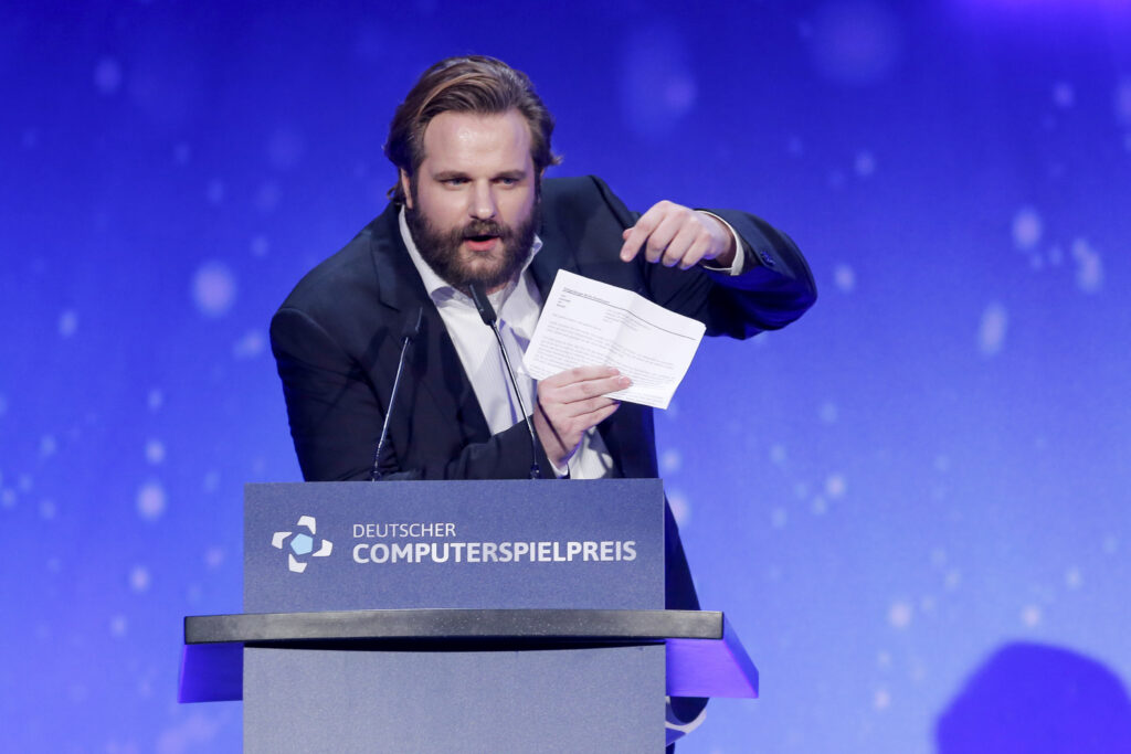 Erik Ranke steht am Rednerpult Deutscher Computerspielpreis und hält einen weißen Zettel in der Hand