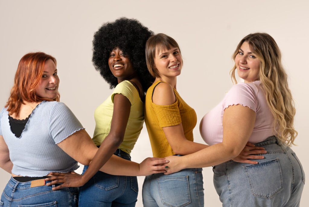 Vier Frauen stehen mit dem Rücken zu einer Wand und drehen ihre Köpfe um. Zwei sind auffallend dick, eine andere ist Afrikanerin