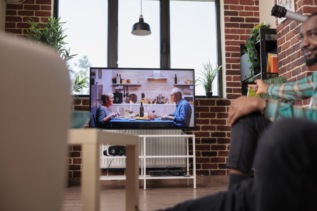 Fernseher im Hintergrund und ein Mann der am Boden sitzt und auf den Fernseher deutet