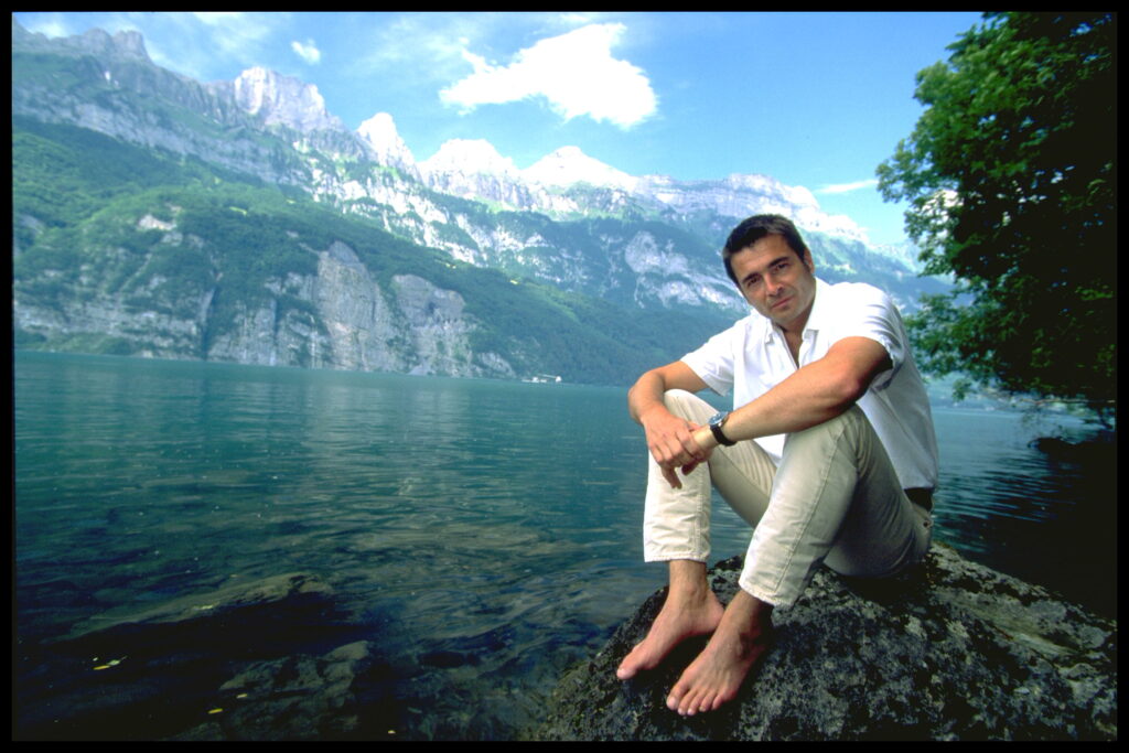 Stefan Gubser sitzt mit nackten Füssen auf einem Stein an einem See. Dahinter eine Bergkette