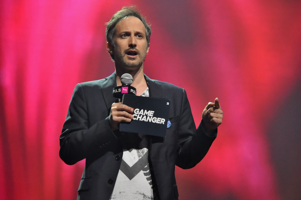 Michael Ostrowski mit schwarzem Sakko und Mikrofon von Puls4 hält Game Changer Karte in einer Hand