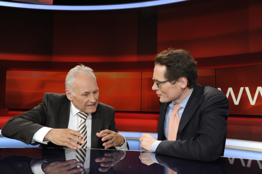 Roger Köppel sitzt neben einem Mann an einem Moderationspult