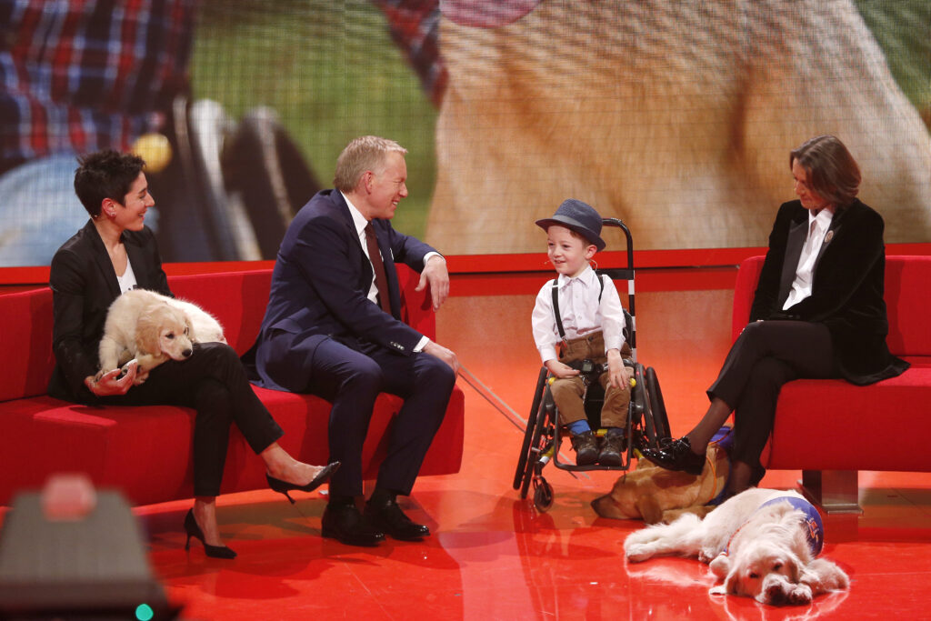 Dunja Hayali mit einem Hund auf ihrem Schoss neben einem Mann auf einem roten Sofa. Weiters ein Kind im Rollstuhl, zwei weitere Hunde und eine Frau
