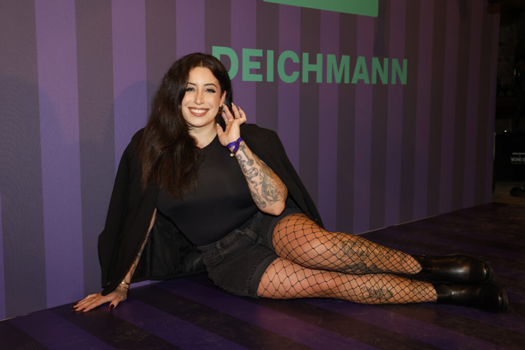 Jolina Mennen mit schwarzem Outfit sitzt vor einem Werbeplakat von Deichmann